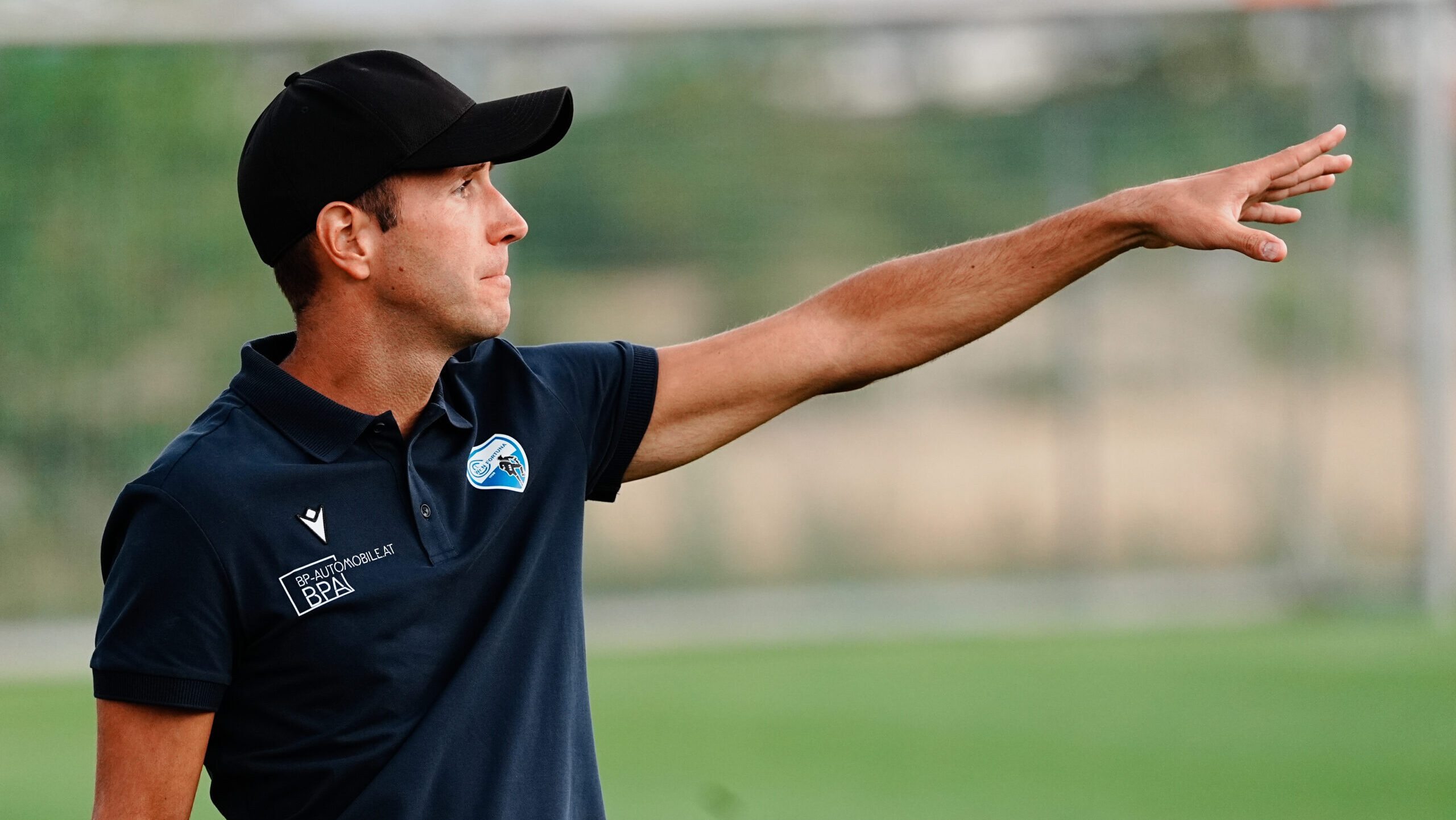 Trainer mit schwarzer Kappe zeigt am Fußballplatz nach rechts