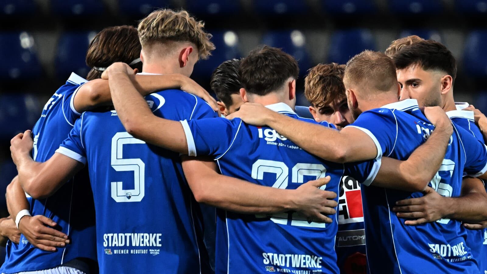 Fußballspieler im blauen Dress mit Nummern 5 und 22 umarmen sich in einer Huddle.
