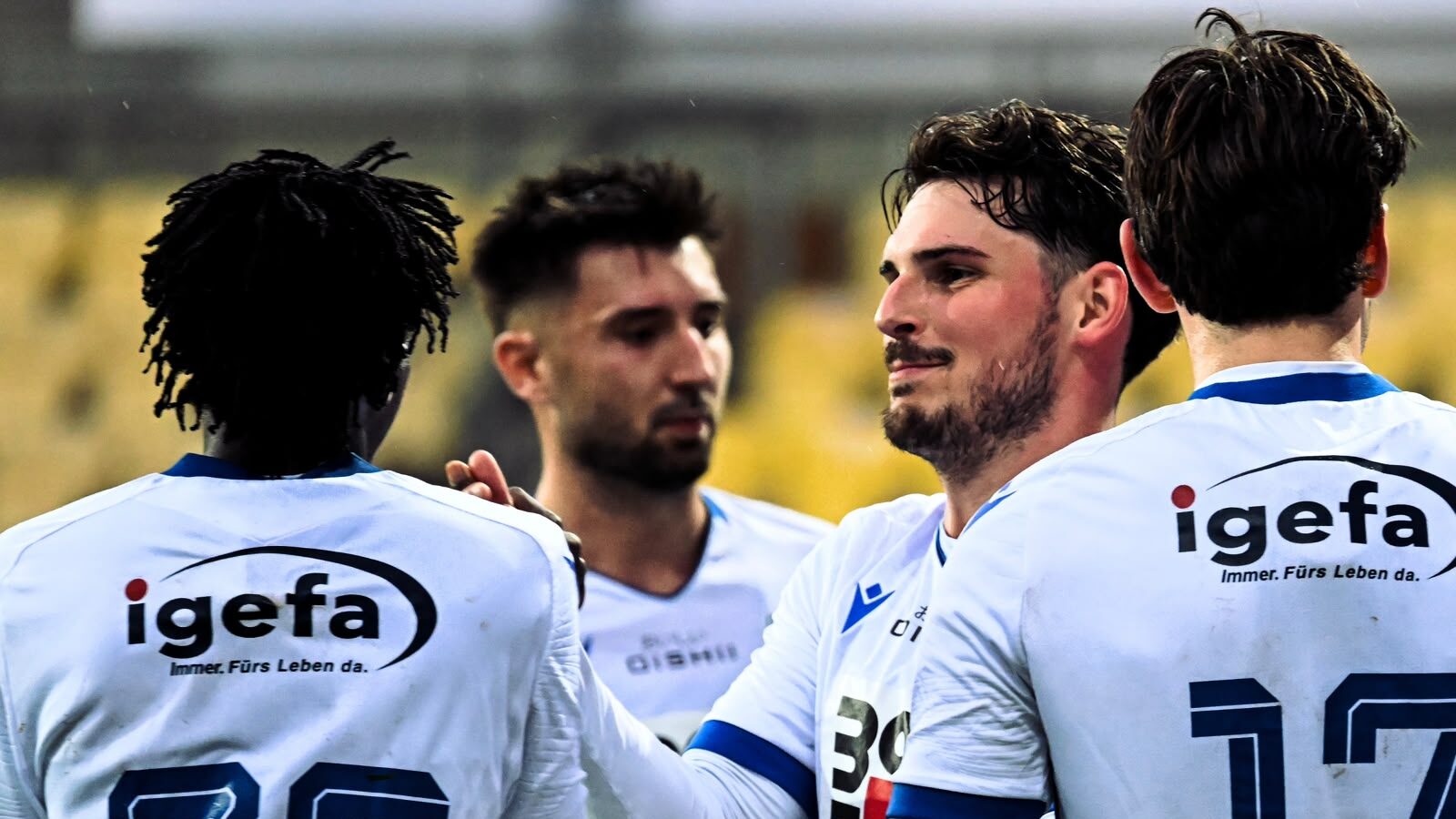 Fußballer mit Dreadlocks und Bart im Jubel, mit Spielern im Hintergrund.